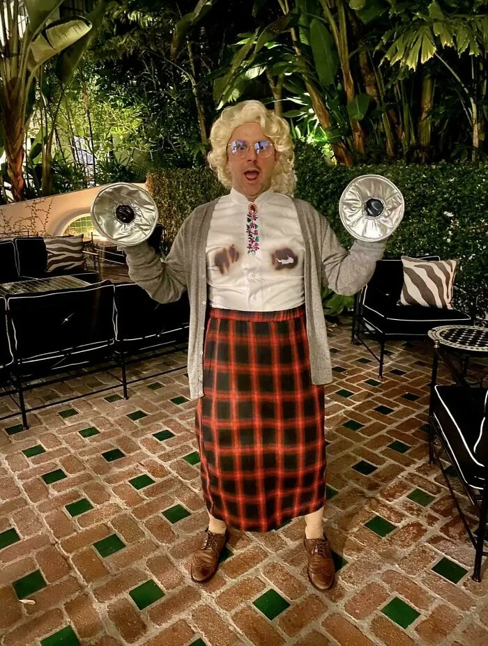 Person wearing a genius Halloween costume with a blonde wig, glasses, gray cardigan, and red plaid skirt at a party outdoors.
