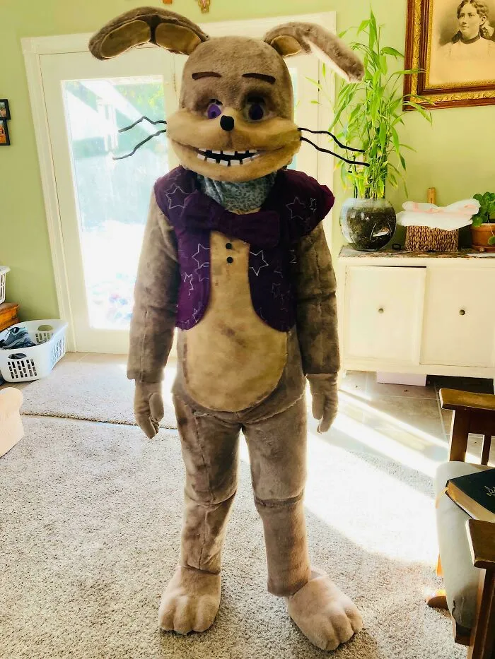 Person in a genius Halloween costume resembling a large plush rabbit with a purple vest, standing in a living room.
