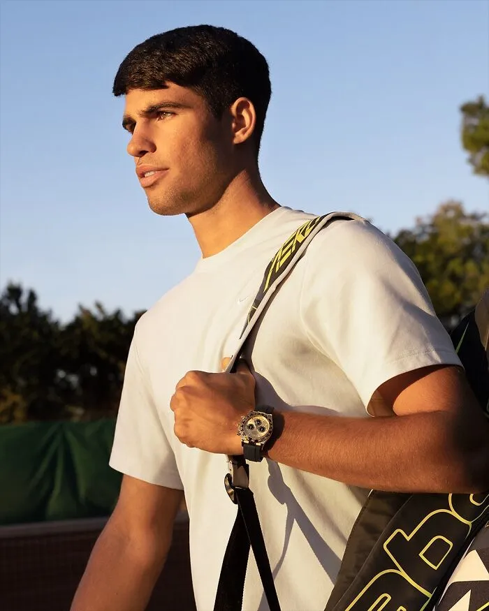 Carlos Alcaraz outdoors wearing a white shirt and watch, carrying a sports bag, representing the most handsome man in the world 2025.
