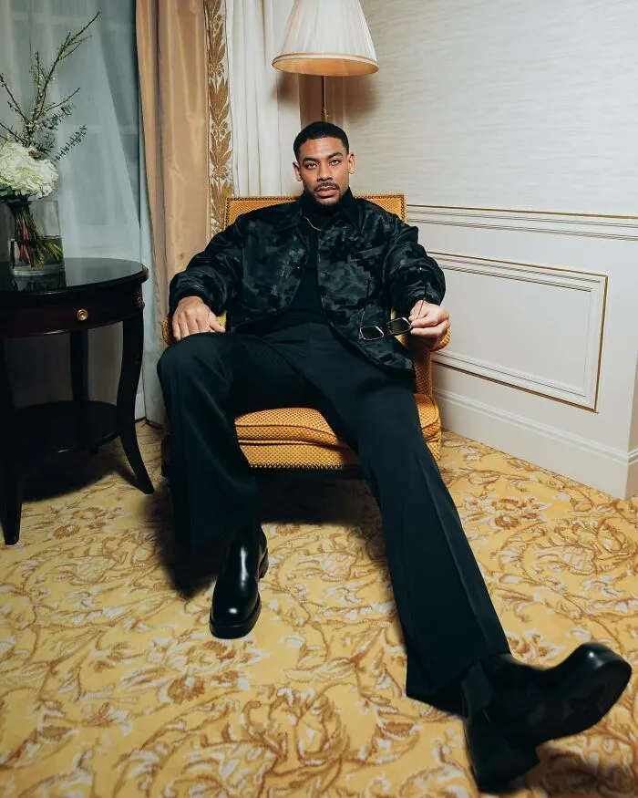 Aaron Pierre sitting confidently in black outfit holding sunglasses indoors, representing most handsome man in the world 2025.