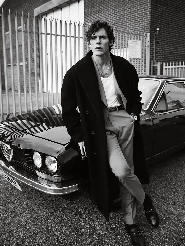 Sam Claflin in a long coat leaning on a vintage car, embodying classic charm and handsome man fashion 2025.