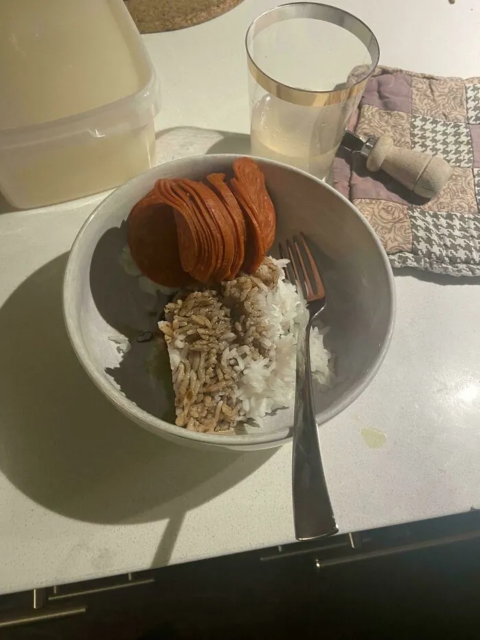 Bowl with white rice, slices of pepperoni, and a dark sauce, accompanied by a glass of water on a kitchen counter unholy food pics.