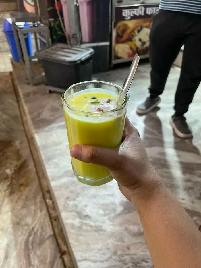 Hand holding a glass of yellow drink with spoon in a local food setting, highlighting foods not popular in their country.