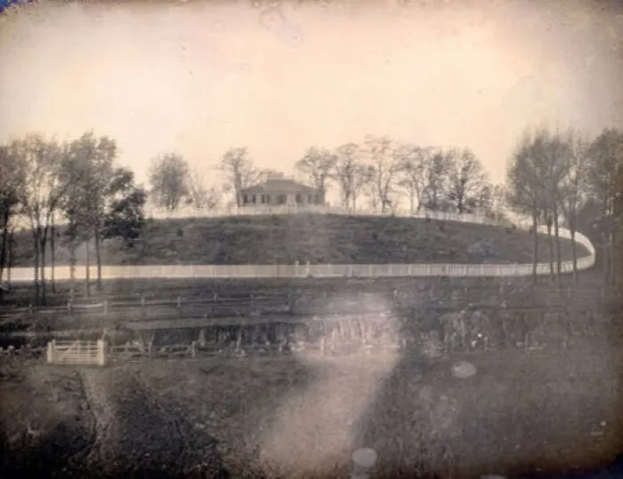 Early photography image showing a house on a hill surrounded by trees and fences from the time photography was new.