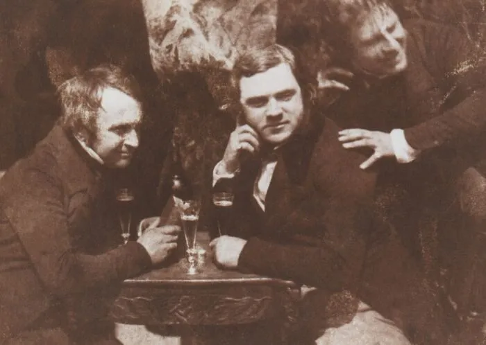 Three men in vintage clothing around a table with glasses, captured in an early photography era historic photo.