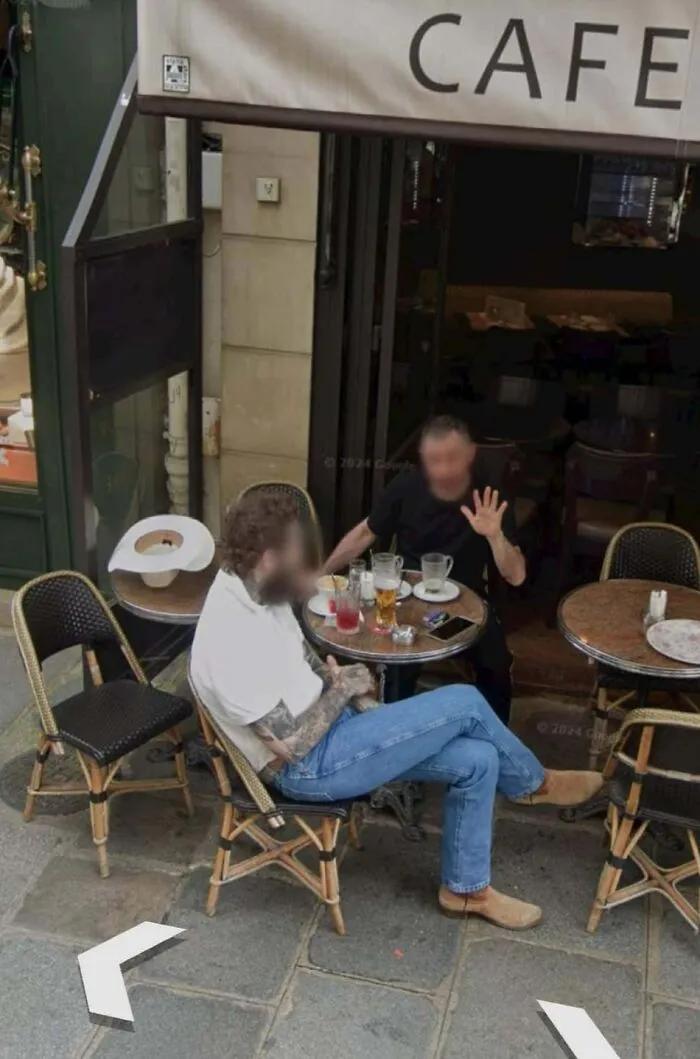 Two men sitting outside a café with drinks, captured unexpectedly in a Google Earth and Maps street view image.