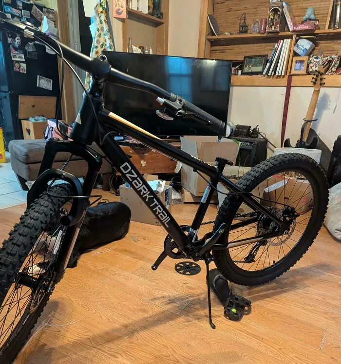 Bicicleta de montaña negra Ozark Trail en sala desordenada como ejemplo de compras en línea que salen mal.