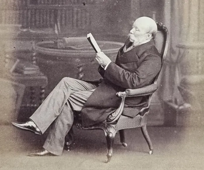 Elderly man in vintage attire sitting in an ornate chair reading a book about famous mysterious imaginary worlds.