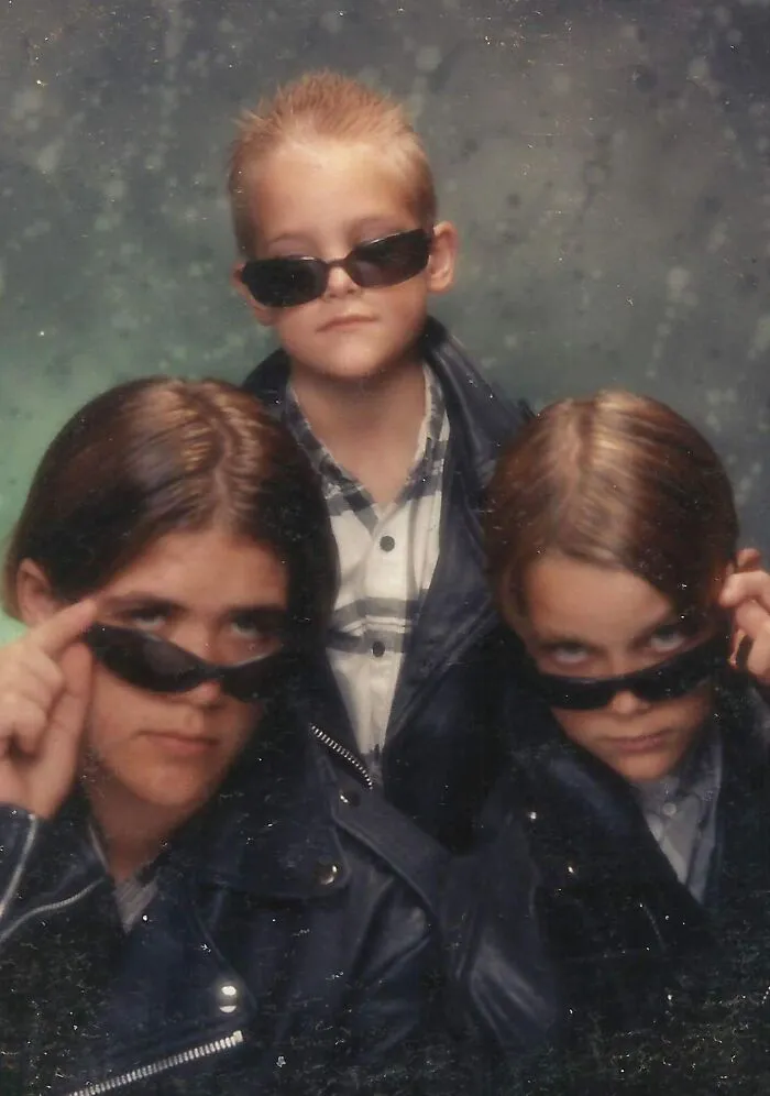 Tres hermanos con gafas oscuras y chaquetas de cuero posando para una foto familiar incómoda y divertida.