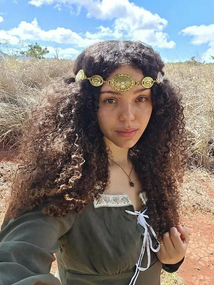 Young woman with curly hair wearing a vintage dress and gold headpiece in a natural field, showcasing cottage core aesthetics.