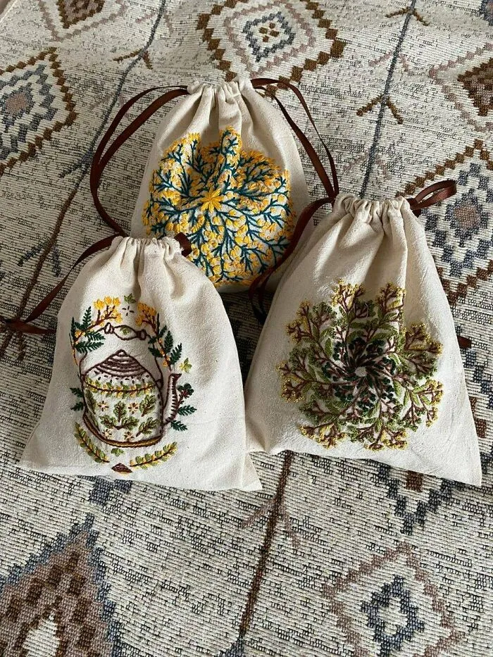 Three drawstring bags with floral and teapot embroidery, showcasing charming cottage core aesthetics on a patterned fabric background.
