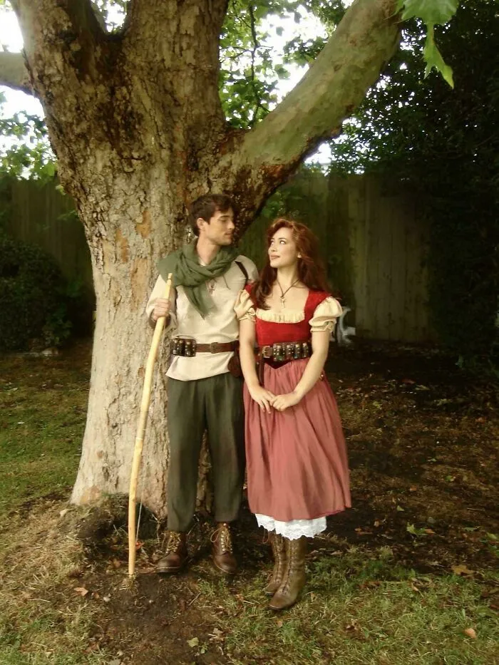 Couple dressed in cottage core aesthetics outfits standing by a large tree in a whimsical outdoor setting.