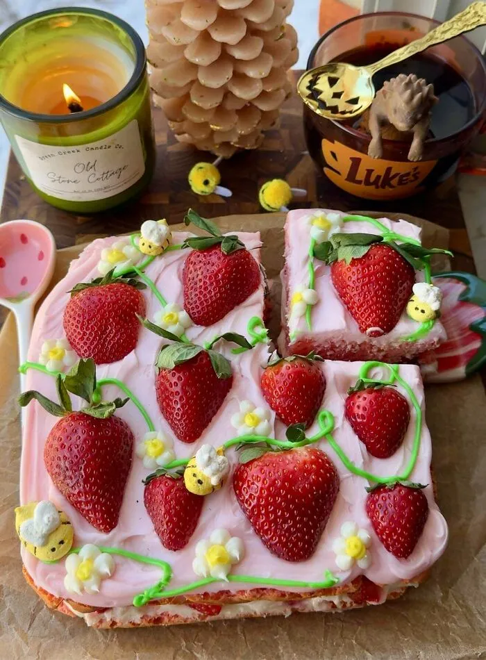Square strawberry cake with pink frosting decorated with bees and flowers, surrounded by cottage core aesthetics items on a wooden board.