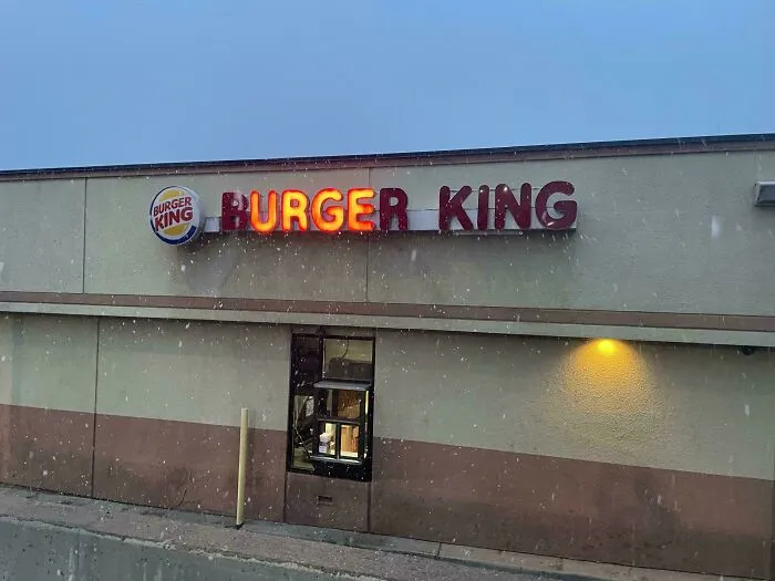 Burger King LED sign with partially lit letters on building exterior during snowfall at dusk.