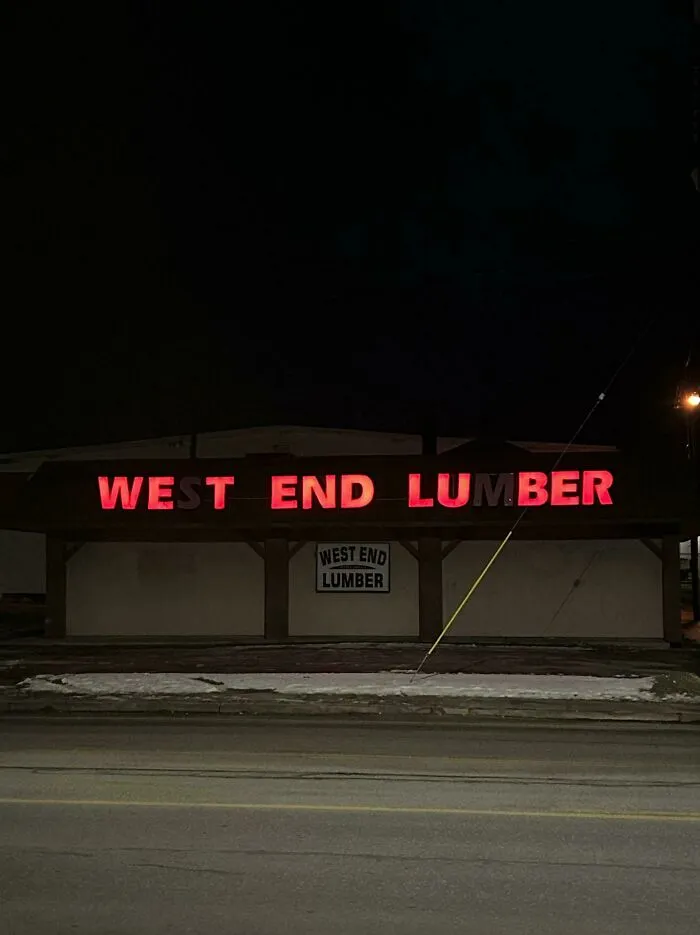 Red LED sign displaying West End Lumber on a building at night along a snowy roadside.