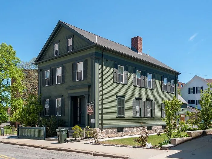 Historic green haunted house with black shutters under clear blue sky, a spooky destination for Halloween visitors.