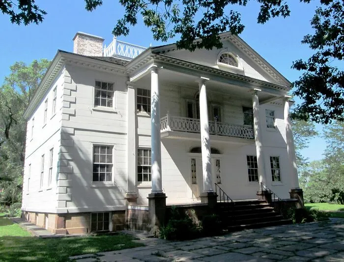 Historic white mansion with large pillars and balcony, surrounded by trees, one of the spooky destinations to visit this Halloween.