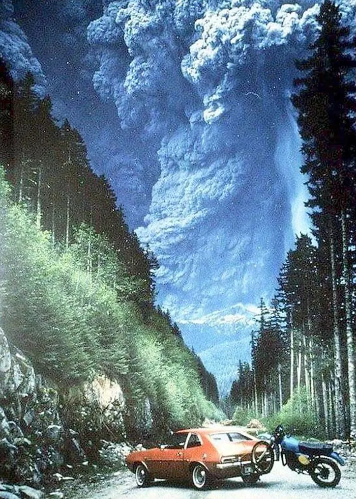 Erupción volcánica espectacular en los años 1980 con coche y motocicleta detenidos en carretera rodeada de bosque.