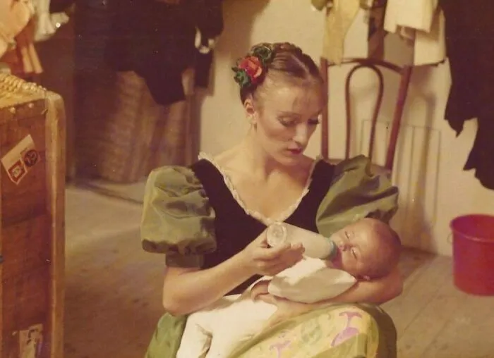 Mujer con vestido estilo años 80 alimenta a un bebé con biberón, mostrando moda y vida cotidiana en los 1980s.