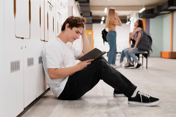 Teen boy sitting alone by lockers reading a book while popular kids talk in the background in school hallway outcasts