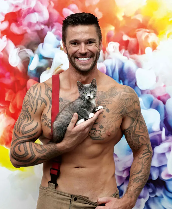Shirtless Australian firefighter with tattoos holding a gray kitten against a colorful abstract background for calendar shoot