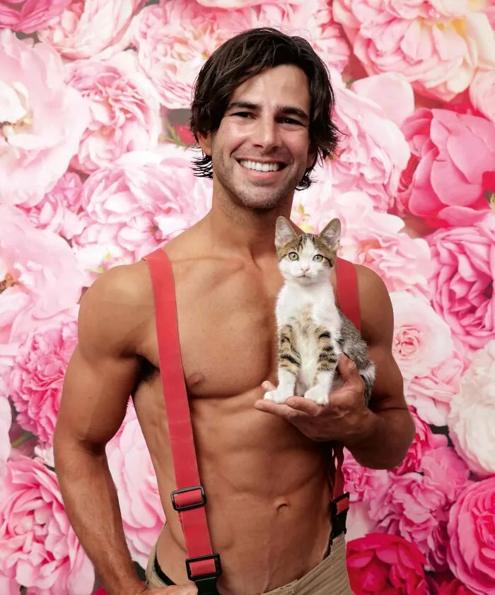 Shirtless Australian firefighter wearing red suspenders holding a kitten against a pink floral backdrop.
