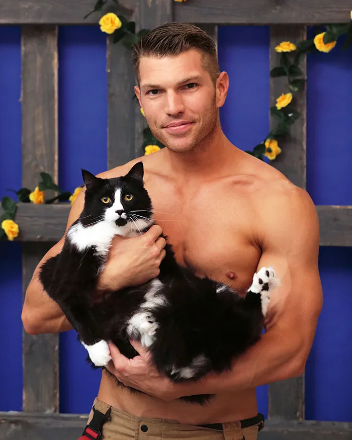 Shirtless Australian firefighter holding a black and white kitten against a blue backdrop with yellow flowers.