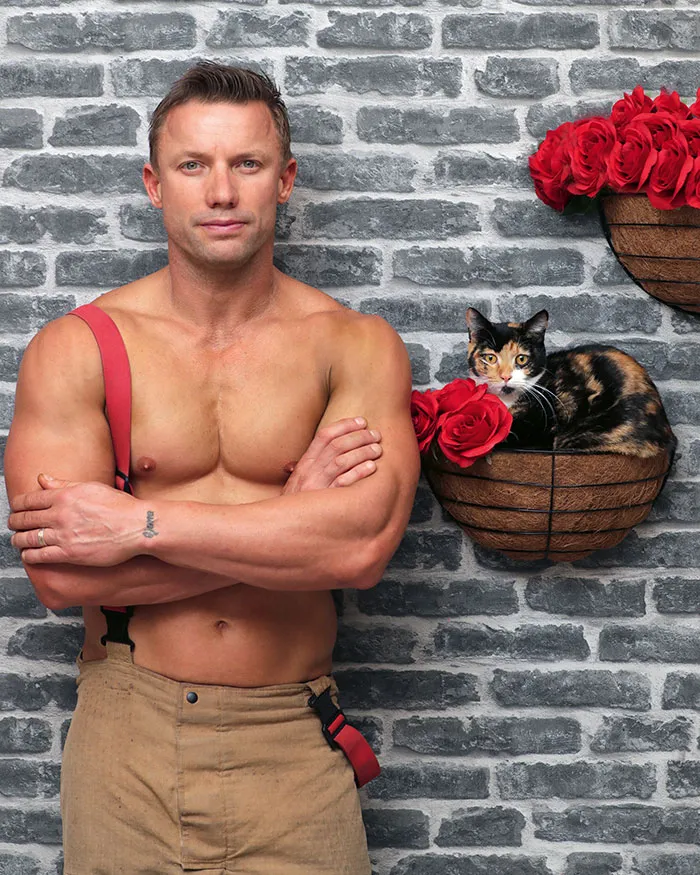 Shirtless Australian firefighter posing with a calico kitten and red roses in a basket against a gray brick wall.