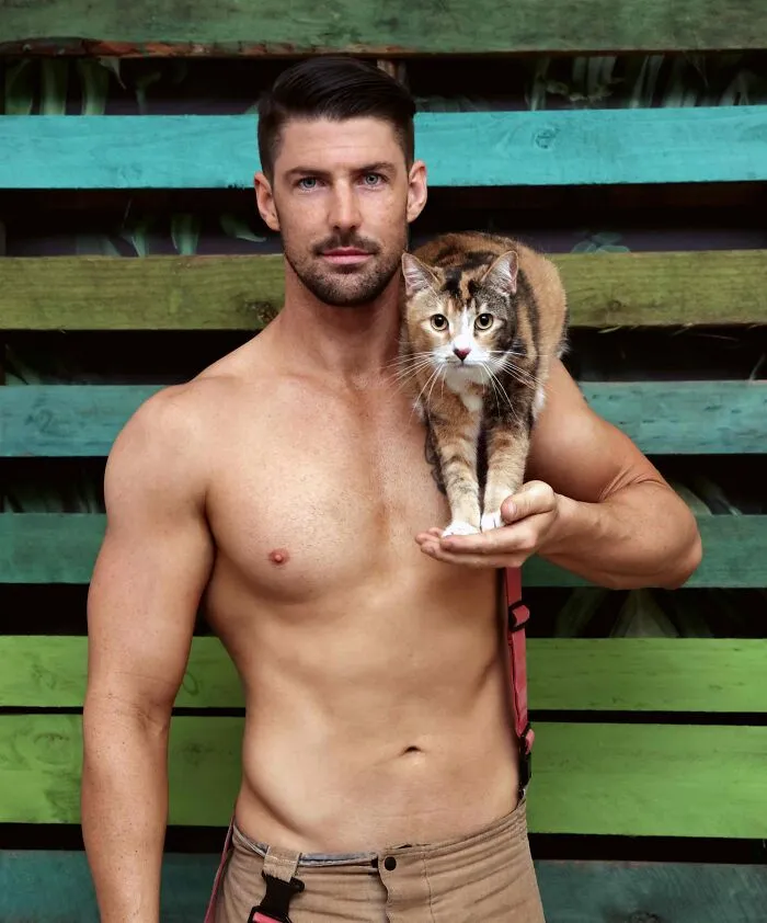 Shirtless firefighter holding a kitten on his arm posing against a colorful wooden background for Australian firefighters calendar.