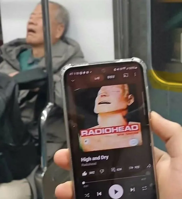 Person holding phone showing Radiohead song playing, with sleeping elderly man in the background on public transit.
