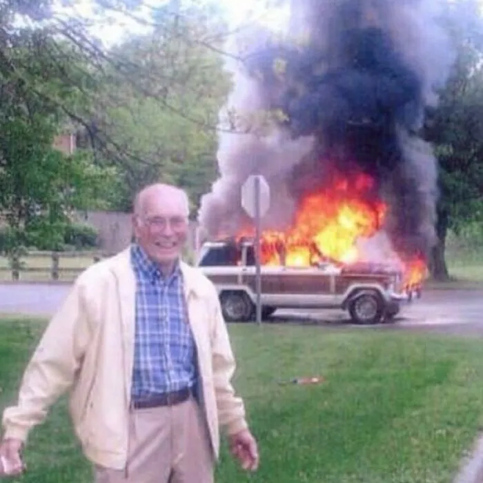 Hombre mayor sonriente frente a un coche en llamas, imagen extraña y bizarra con fuego y humo intenso en fondo.