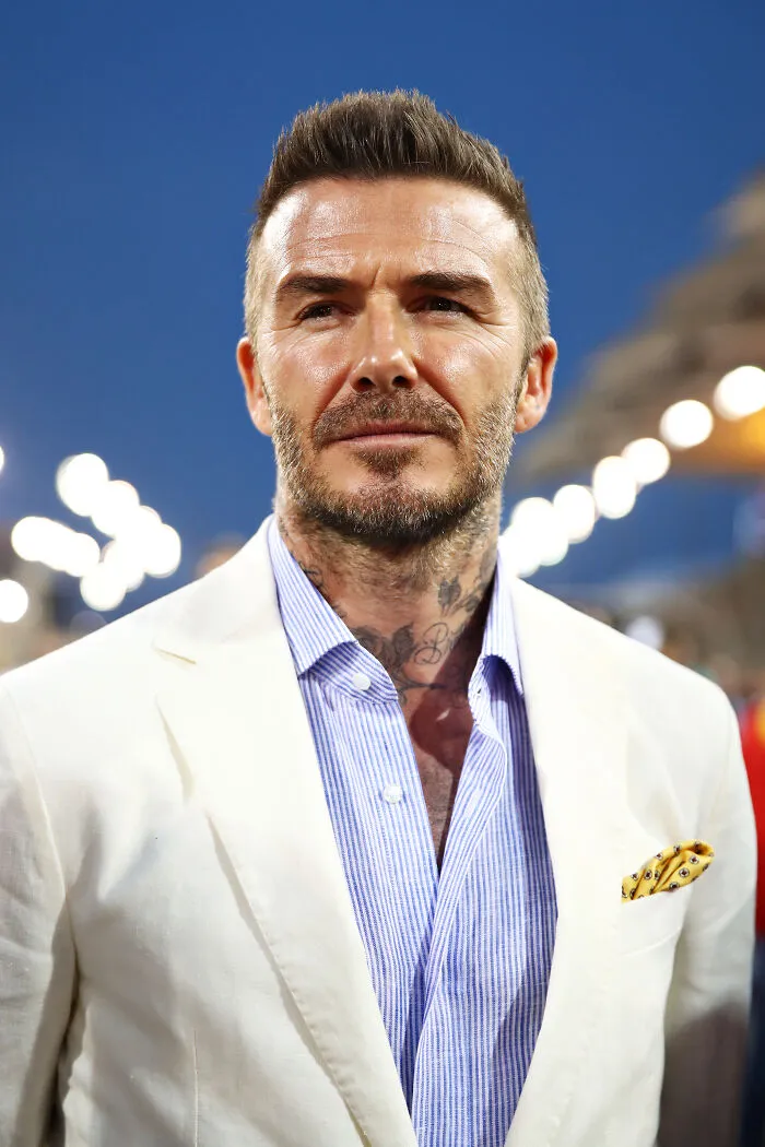 Man with styled hair and beard wearing a white blazer and striped shirt at an evening event, most handsome man in the world.