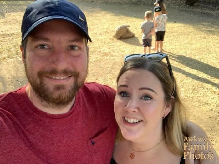 Pareja sonriente en selfie con niños y oveja de fondo, foto familiar incómoda icónica al aire libre en días soleados.
