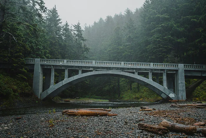 Concrete arch bridge over a rocky riverbed surrounded by dense forest, evoking spooky urban legends atmosphere.