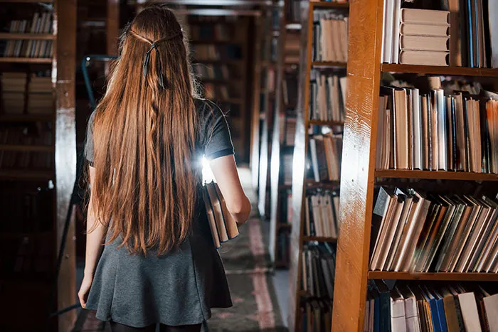 Young woman with long hair holding books, exploring a dimly lit library, evoking spooky urban legends atmosphere.