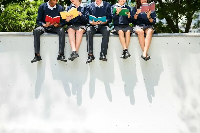 Group of popular kids in school uniforms sitting on a wall reading books, showing shocking school behaviors that caused outcasts.