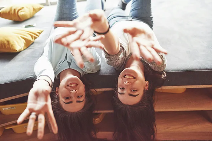 Two smiling young women lying upside down on a couch reaching out, representing famous supermodels in a casual setting.