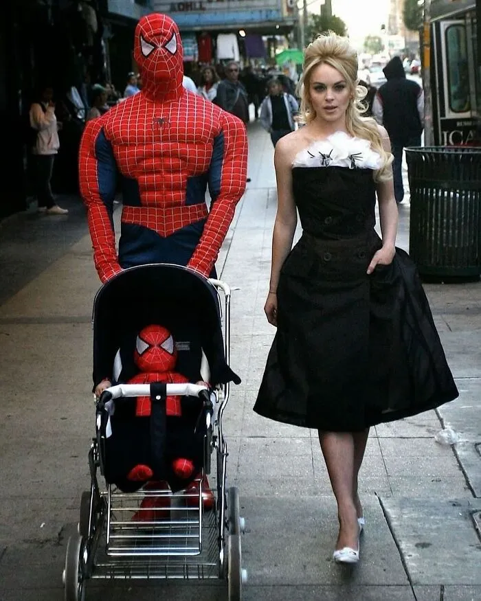 Spiderman in costume pushing a stroller with a child in matching outfit, walking beside a woman in a black dress on a city sidewalk.