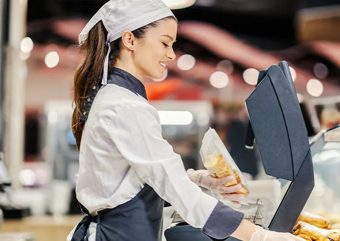 Smiling food service worker wearing uniform and gloves, handling packaged food while operating a cash register in a busy setting.