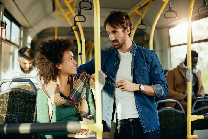 Two multilingual people catching gossip in secret languages while standing and sitting on a public bus during daytime.