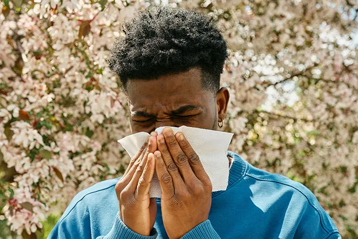 Young man in a blue sweater sneezing into a tissue outdoors with blooming trees, illustrating red flags in relationships concept.