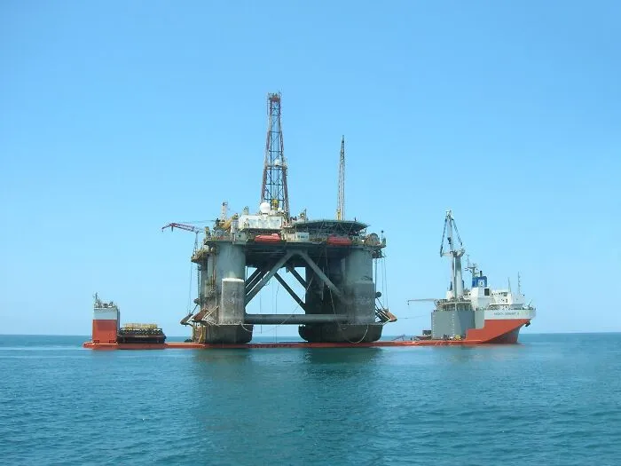 Offshore oil rig platform being transported by cargo ship on calm ocean under clear blue sky, disturbing Wikipedia pages.