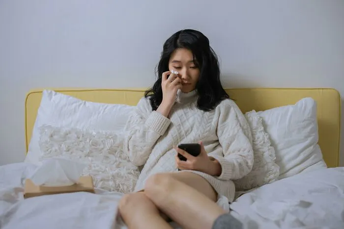 Woman sitting on bed wiping tears, feeling upset while using phone, illustrating the impact of living next to horrible people.
