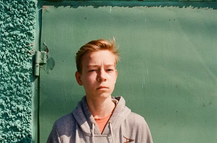 Teenage boy standing against a green textured wall, looking serious, reflecting dark secrets that could affect lives.