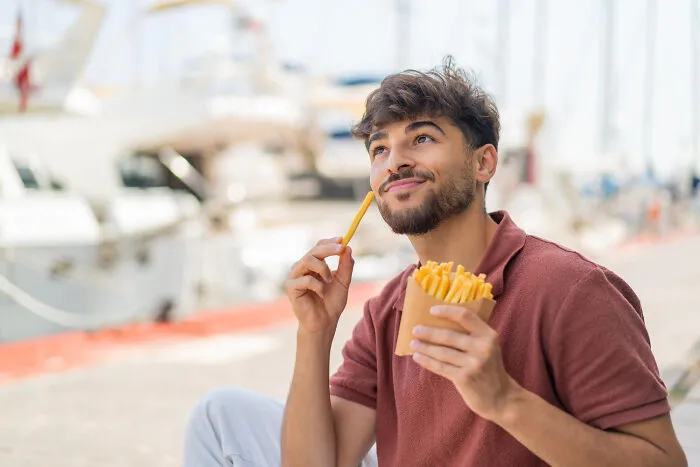 Young man enjoying fries outdoors by the marina, reflecting on little lies that backfired in everyday life.