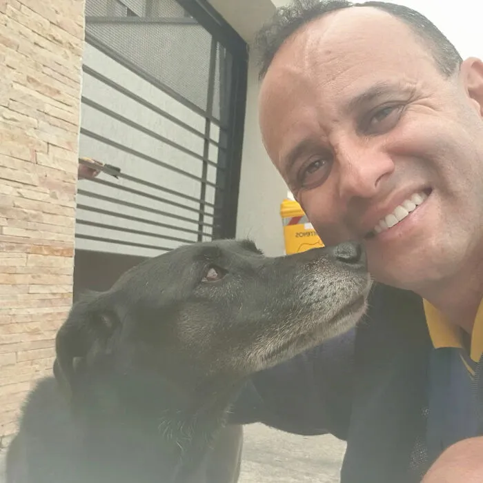 Mailman smiling while a black dog affectionately nuzzles his face during a friendly moment on his route.