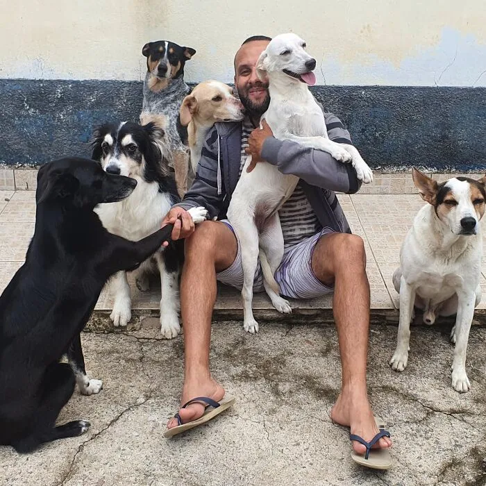 Mailman happily surrounded by friendly dogs on his route, sitting and bonding with them outdoors.