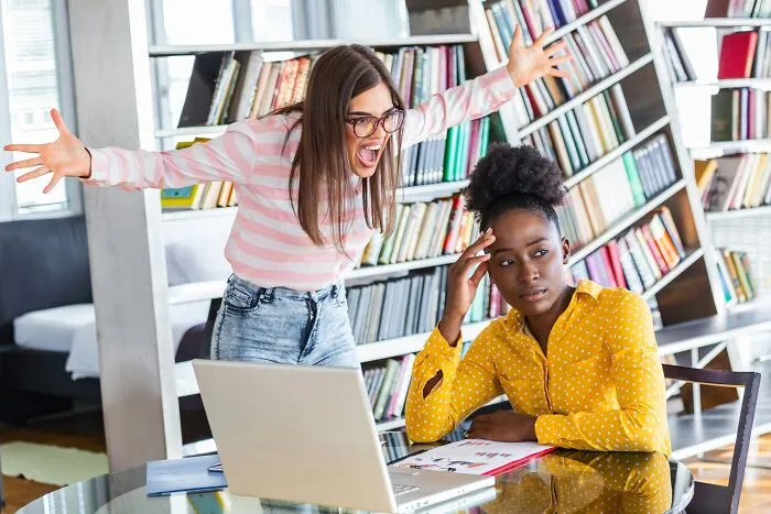 Two women in a library showing toxic relationship traits, one frustrated and yelling while the other looks distressed at a laptop.