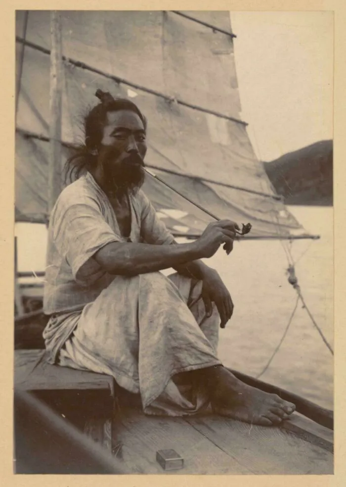 Un marinero coreano descansa de su trabajo transportando carga, sentado a la sombra de la vela, fumando en su larga pipa de bambú. Viste ropa fresca de cáñamo. Alrededor de 1904.