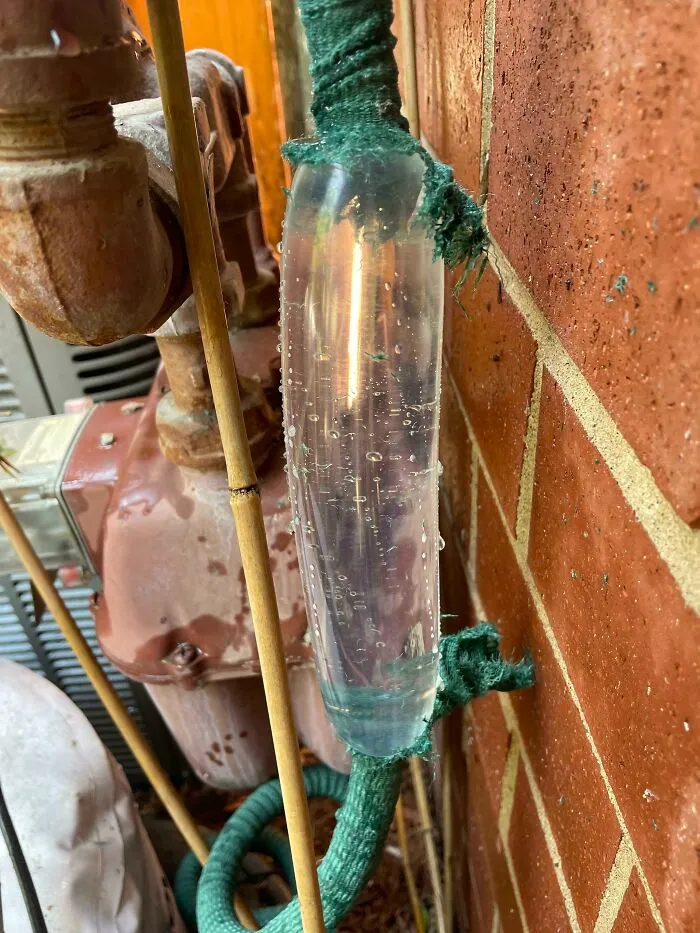 Close-up view showing the insides of a transparent water hose with bubbles trapped inside near rusty machinery and brick wall.